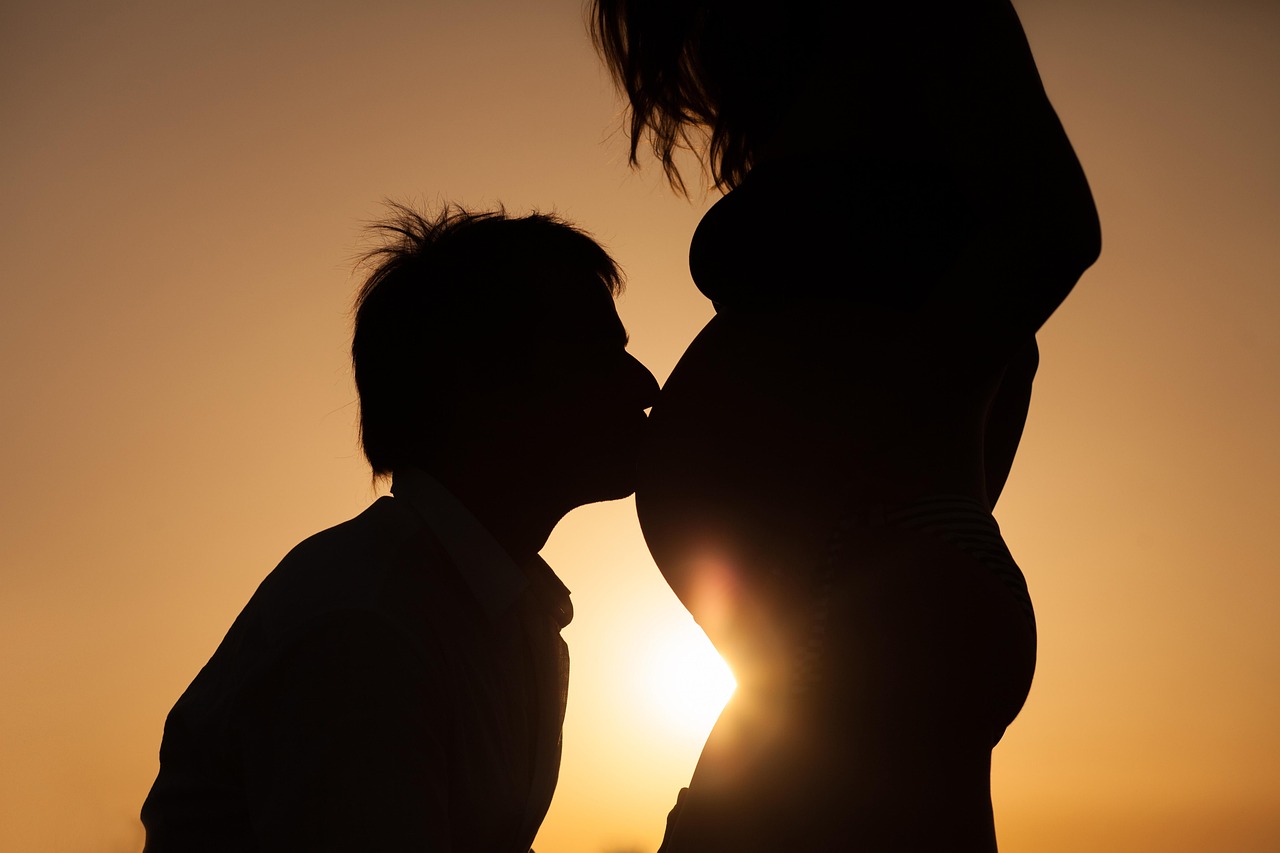 Mujer embarazada con su pareja besándole la barriga al atardecer, con una puesta de sol en el fondo.